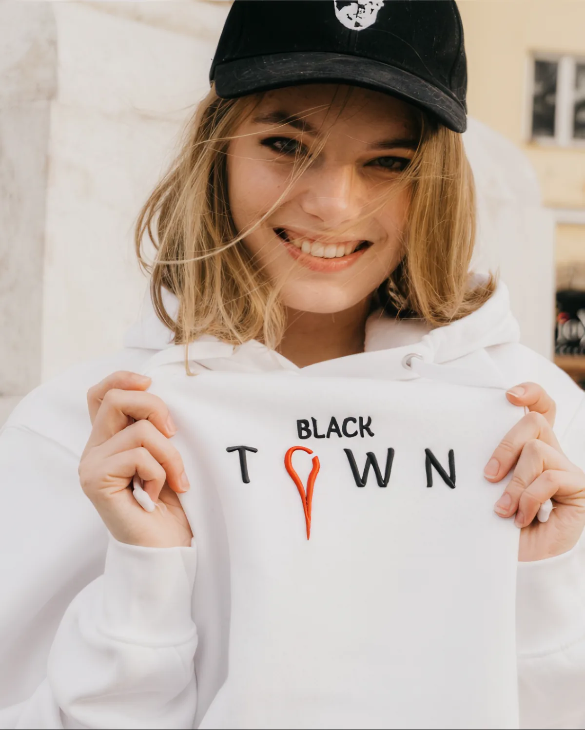 Usměvavá dívka v černé kšiltovce drží bílou mikinu s výšivkou Black Town