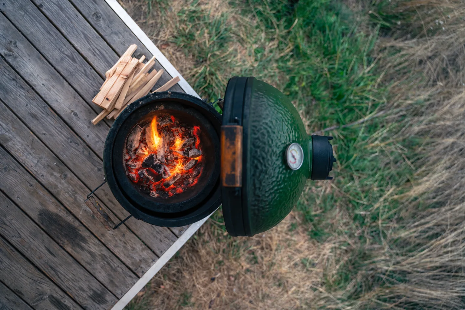 Pohled shora na otevřený zelený kamado gril s rozžhaveným uhlím vedle třísek na terase