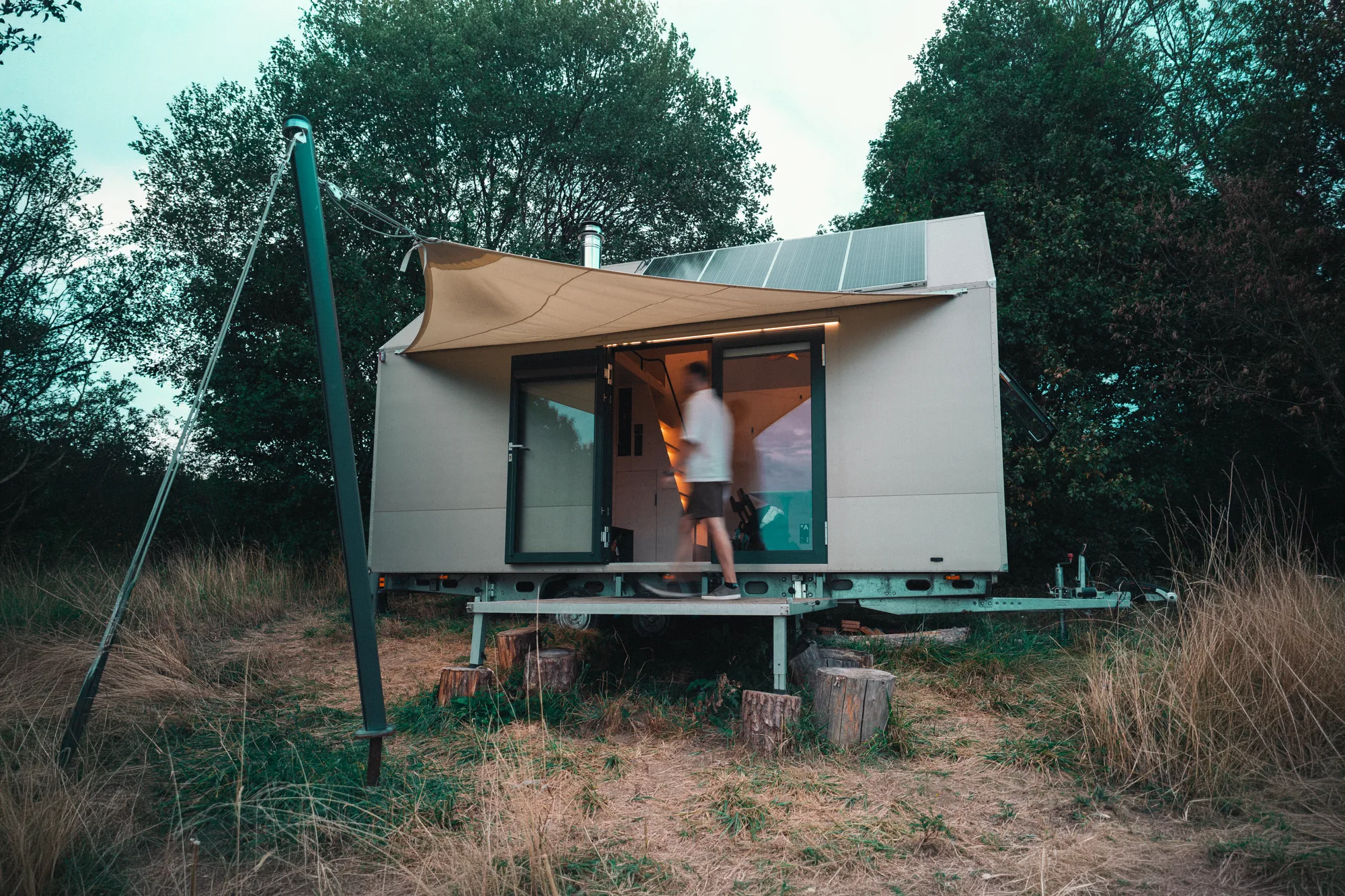 Tiny house za soumraku s rozsvíceným interiérem a solárními panely, kolem prochází rozmazaná postava